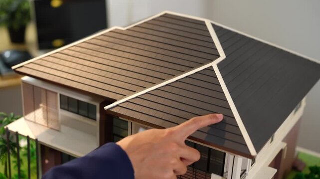 Close view of a house model with a hand pointing at the roof, showing home design, inspection, real estate planning, or property investment in an indoor setting.