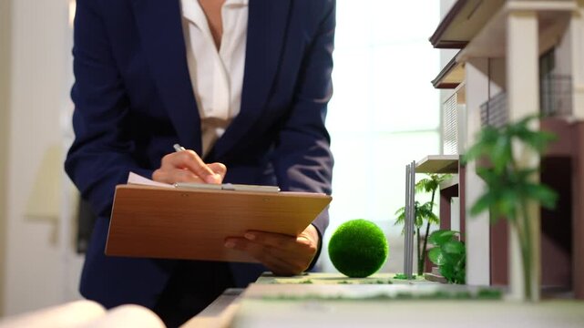 Businesswoman writing on a clipboard beside a modern house model, suggesting property inspection, real estate planning, home sales, or architectural review in a bright office.