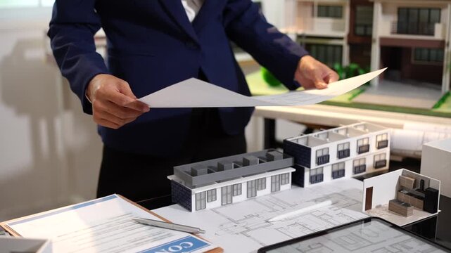 Real estate agent or architect reviewing house plans beside a scale model home and apartment building in an office, showing property planning, design, and development work.