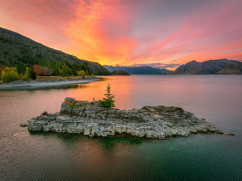 New Zealand - 24 April 2022: View of a rocky island with a single tree in Lake Wanaka at sunset, with vibrant orange and pink clouds reflecting on the water and mountains in the distance.
