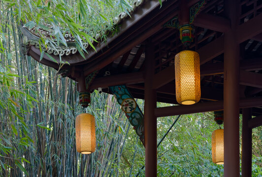 A serene scene featuring glowing yellow lanterns hanging from an ornate traditional pavilion roof amidst a lush bamboo forest in WangJiangLou park. The composition highlights the harmony between
