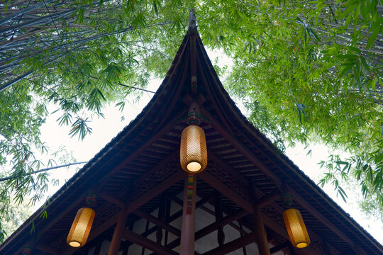 A low-angle view of a traditional Chinese pavilion roof featuring intricate woodwork and glowing lanterns nestled among dense green bamboo foliage. The scene captures a tranquil, cultural atmosphere