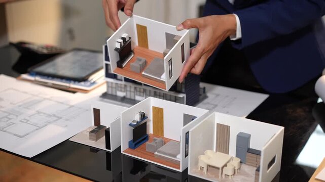 Architect or designer arranging a modern house model on a desk with floor plans, showing home layout, interior design, planning, and real estate development.
