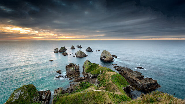 New Zealand - 18 January 2019: View of rugged coastal cliffs and rocky islets rising from the sea under a dramatic cloudy sky at sunset.