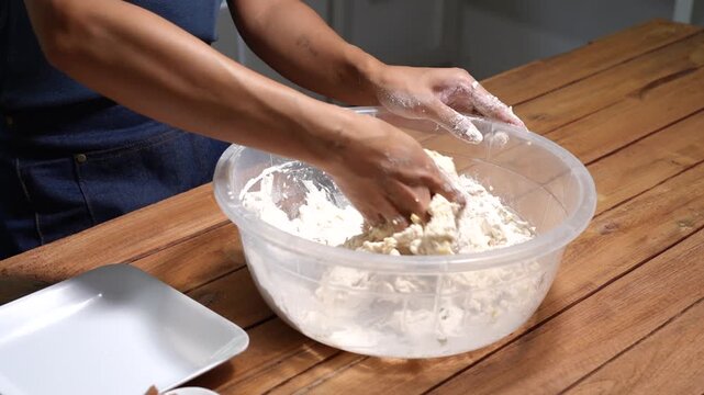 Close-up of hands mixing flour dough in a clear plastic bowl on a wooden table, showing home baking, cooking process, and fresh food preparation in a simple kitchen setting.