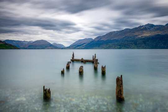 New Zealand - 26 March 2021: View of weathered wooden posts of an old jetty rising from the calm, long-exposure waters of Lake Wakatipu, set against dramatic mountains and a cloudy sky.