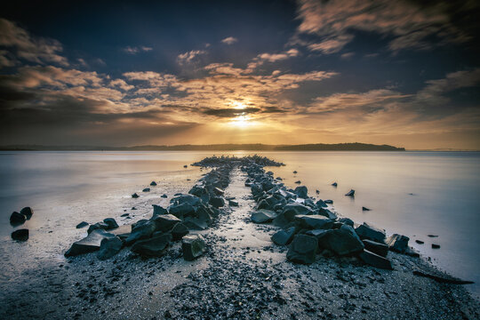 New Zealand - 10 June 2021: View of a rocky breakwater extending into the calm sea under a dramatic sunset sky.