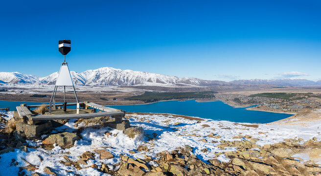 New Zealand - 09 July 2021: View of the trig station on Mount John overlooking the turquoise waters of Lake Tekapo and the snow-capped Southern Alps under a clear blue sky.