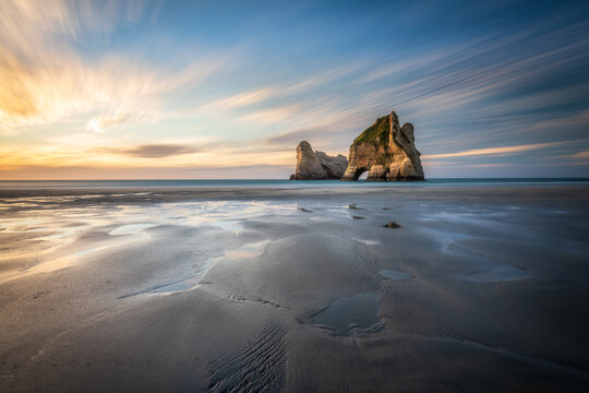 New Zealand - 13 February 2021: View of the Archway Islands at Wharariki Beach, featuring dramatic rock formations, tidal pools on the sandy shore, and a vibrant sunset sky.