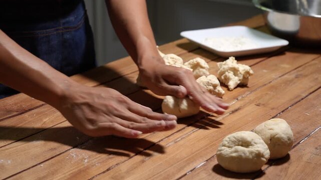 Close view of hands kneading dough on a wooden table in a kitchen, with dough pieces and baking tools nearby, showing homemade bread preparation and warm rustic cooking.