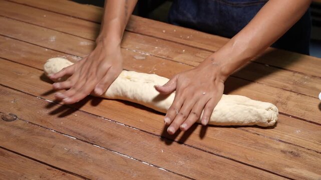 Close view of hands rolling bread dough on a rustic wooden table with flour, showing home baking, cooking process, and handmade food preparation in a warm kitchen setting.