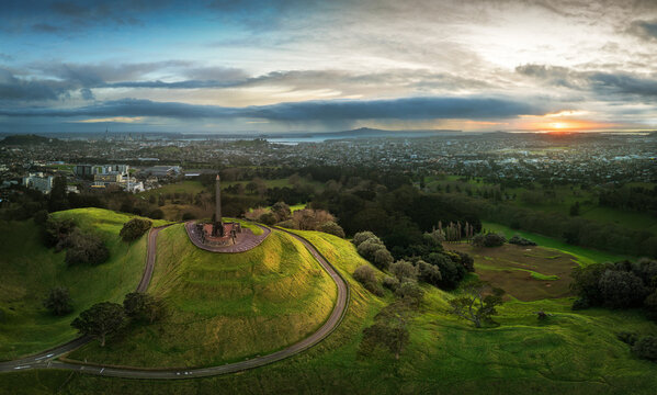 Auckland, New Zealand - 18 August 2026: View of the One Tree Hill monument on a grassy volcanic cone with a city skyline and sunset in the background.