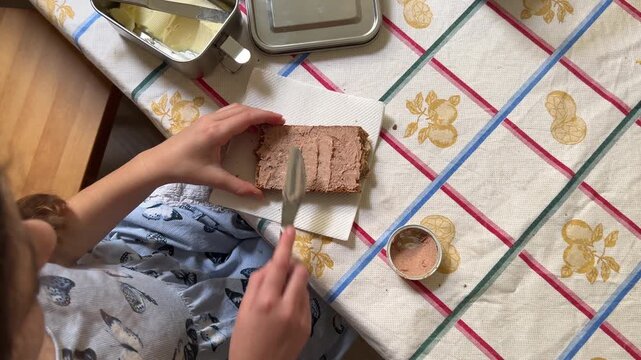 Horizontal top-down shot of a girl preparing her snack, spreading pate on a slice of bread resting on a napkin using a knife.