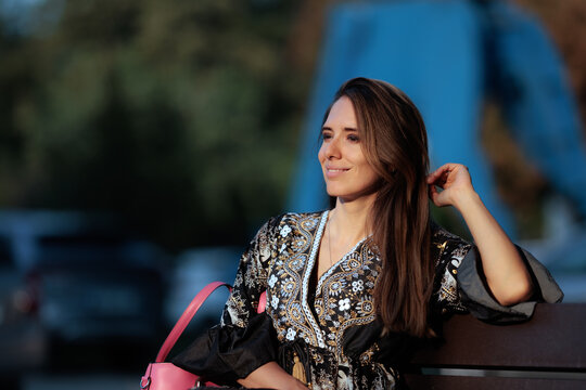 Beautiful Smiling Woman Sitting On Bench Running Hand Through Her Hair. Confident and elegant young lady sitting outdoors while gently touching her brown hair
