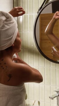 Vertical shot of Hispanic young woman with towel on head spraying deodorant under armpit in bathroom. Clip is perfect for personal hygiene, body care, and morning routine advertisements