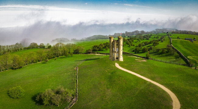 Broadway, England - 22 April 2023: View of Broadway Tower surrounded by lush green fields, stone walls, and rolling hills under a cloudy sky.
