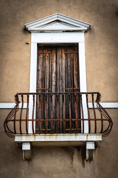 04 October 2022: View of a classic window with weathered wooden shutters, a white stone frame, and an ornate wrought-iron balcony on a textured beige stucco wall.