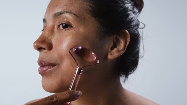 Portrait of young Hispanic woman massaging her face with rose quartz roller. Ideal for promoting skincare products, anti-aging routines, and self-care wellness campaigns