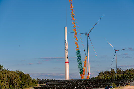Construction of wind turbine with heavy mobile crane and solar panel farm underneath against clear blue sky, renewable hybrid energy concept