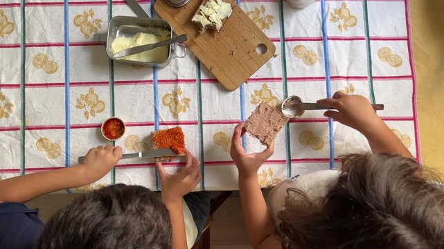 Top-down overhead shot of a girl and a boy preparing their meal, spreading pate and sobrasada on slices of bread with knives on a kitchen table. 