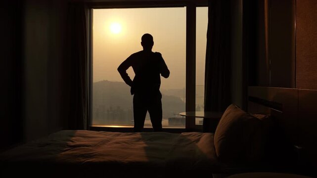 Silhouette of businessman in hotel room getting into focused mindset and preparing for evening meeting, holding his jacket over his shoulder while looking out window at Hong Kong city skyline
