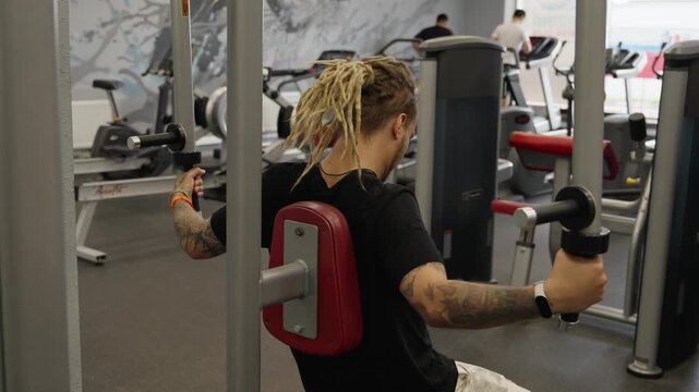 A guy with dreadlocks performs a seated chest fly exercise on a machine at the gym.