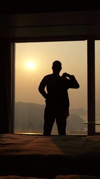 Slow-motion silhouette shot captures confident businessman putting on his jacket in front of floor-to-ceiling window. He stands against bright, glowing sunset, getting ready for evening meeting