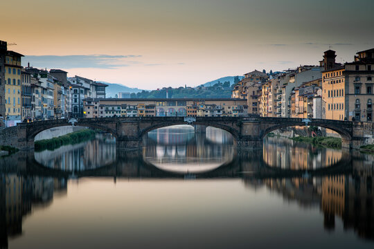 Florence, Italy - 09 May 2023: View of the historic Ponte Vecchio bridge spanning the Arno River at sunset with reflections in the calm water and surrounding medieval buildings.