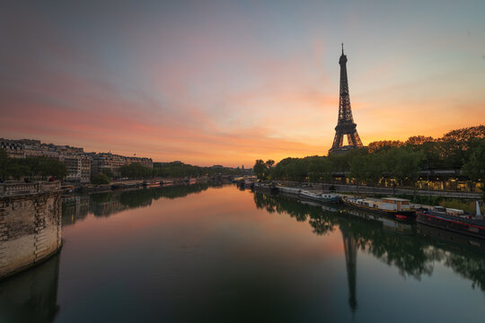 Paris, France - 30 April 2023: View of the Eiffel Tower reflecting in the Seine River at sunset with historic buildings along the banks and a colorful sky.