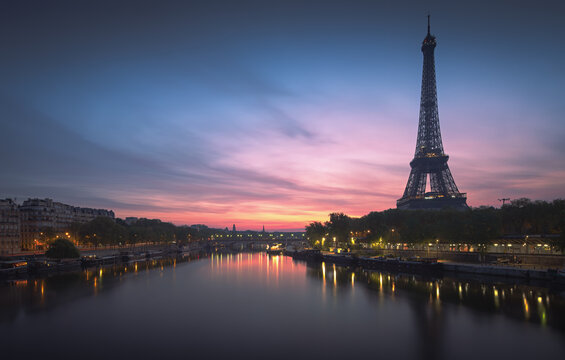 Paris, France - 30 April 2023: View of the Eiffel Tower illuminated at sunset, reflecting in the calm waters of the Seine River with the city skyline in the background.