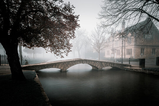 United Kingdom - 22 April 2023: View of a stone bridge over a calm river in a foggy landscape with a historic building in the background.