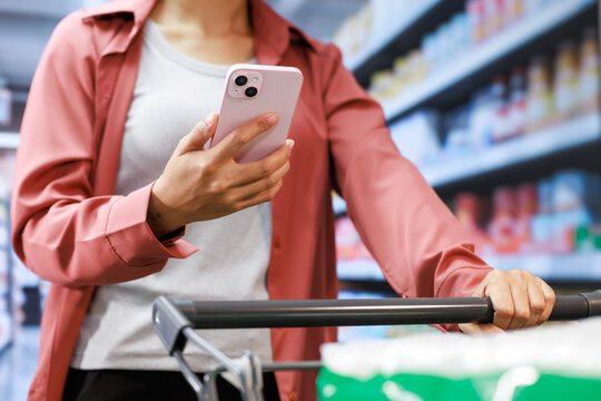 Young woman shopping in a supermarket, holding a smart phone and pushing a shopping cart with shelves blurred in the background