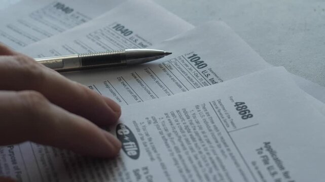 Close-up shot of US tax Form 1040 and Form 4868 with a metallic pen and a person's hand. Person checking official IRS documents for filing income tax returns 