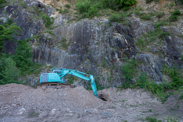 Excavator working at a rocky quarry surrounded by a rugged stone cliff and green vegetation. Industrial construction machinery in a natural landscape, showing mining and excavation activity © Mihail