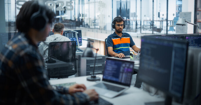 Young Indian Male Programmer in Striped Polo Shirt and Headphones Types Code on a Desktop Computer, Creating Software With a Multiethnic Team of Developers Working on Laptops and Monitors in an Office