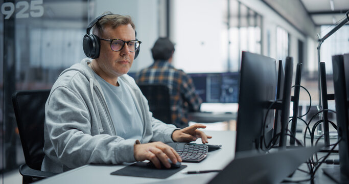 Portrait of a Focused Middle Aged Caucasian Male Software Engineer in Hoodie, Glasses and Headphones Writing Code on a Desktop Computer, Developing Software Beside a Diverse Colleague in an Office.