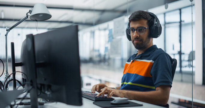 Portrait of a Young Indian Male Software Engineer in Headphones Writing Program Code on a Desktop Computer in a Diverse Office, Developing Applications and Working on Information Technology Projects.