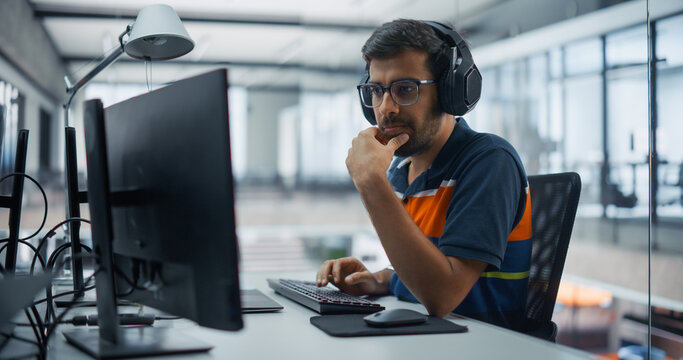 Portrait of a Focused Indian Male Programmer Wearing Headphones, Typing Software Code on a Desktop Computer While Listening to Music, Building Applications With a Team of Engineers in an Office.
