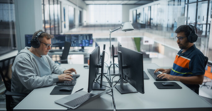 Two Male Software Developers, Caucasian and Indian, in Headphones Listening to Music While Typing Program Code on Desktop Computers at Facing Desks in a Diverse Office, Creating Digital Applications.