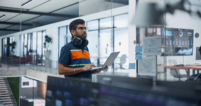 Portrait of Focused Indian Male Software Developer Wearing Headphones Around Neck, Holding Laptop and Studying Sticky Notes and Data Charts While Writing Application Code in a Office of Engineers.