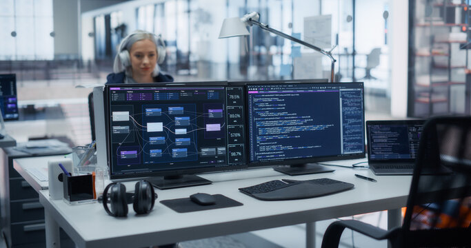 White Female Software Developer Wearing Headphones Writes Program Code Behind Dual Monitors Displaying Data Flow Diagrams and Backend Scripts at a Workstation in an Office of Software Engineers.
