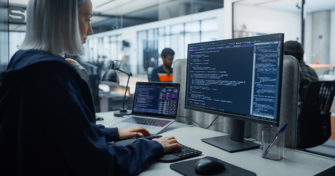 White Female Software Developer Wearing Headphones Types Program Code on Desktop Monitor and Laptop Displaying Scripts at Her Desk in an Office Shared With Diverse Team of Software Engineers.