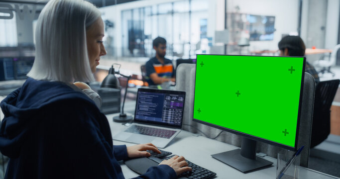 White Female Software Engineer With Headphones Around Neck Types Program Code on a Green Screen Chroma Key Computer While Laptop Displays Scripts at Her Desk in an Office With Development Team.