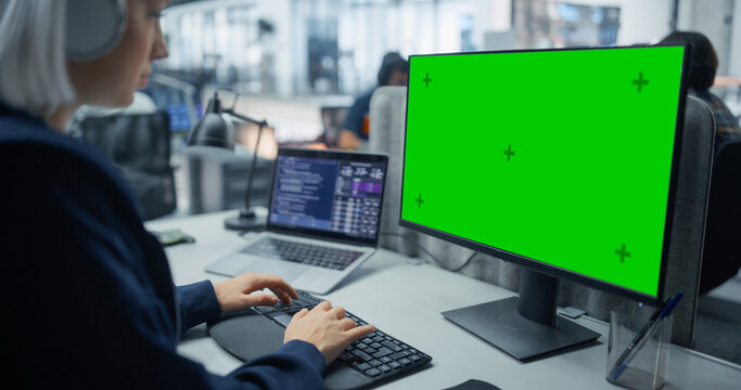 Over the Shoulder View of a White Female Software Developer Wearing Headphones Typing Program Code on a Green Screen Chroma Key Computer and Laptop at Her Desk in an Office With Engineering Team.