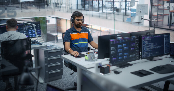 Portrait of a South Asian Male Software Engineer Wearing Headphones Writing Program Code on Dual Monitors Displaying Data Dashboards and Scripts at His Desk in an Office With a Development Team.