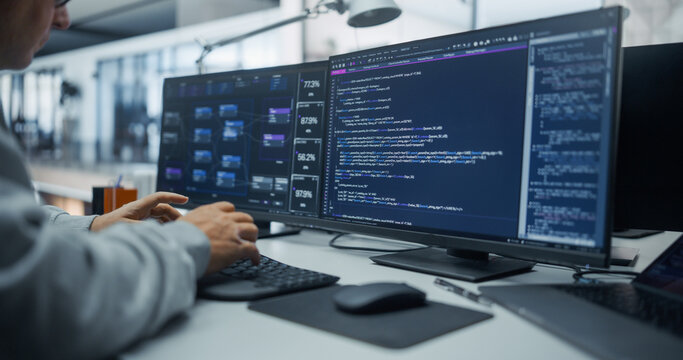 Over the Shoulder View of a White Male Software Developer Writing Database Query Code and Reviewing Data Analytics Dashboards on Dual Monitors at His Desk in an Office With a Diverse Engineering Team.