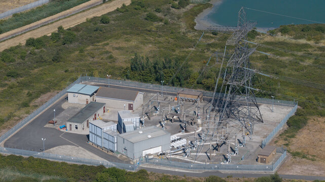 Aerial view of Damhead Creek Power Station showing electrical infrastructure, a large transmission tower, and a fenced compound in the green landscape of Rochester, England, United Kingdom.