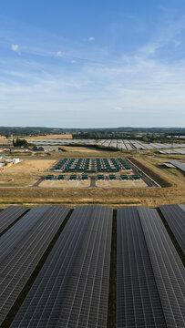Aerial view of a large solar farm featuring rows of photovoltaic panels and battery storage units in a rural landscape under a clear blue sky in Faversham, England, United Kingdom.