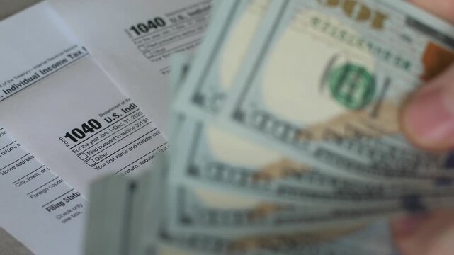 Close-up shot of hands counting hundred-dollar bills over official US Individual Income Tax Return Form 1040. Person counting cash money for tax payment or tax refund process