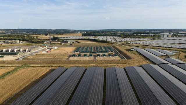 Aerial view of a large-scale solar farm featuring extensive rows of photovoltaic panels, a central battery energy storage system, and surrounding fields in Faversham, England, United Kingdom.
